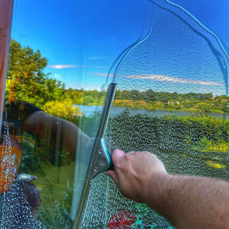 Eine Hand reinigt ein beschlagenes Fenster mit Blick auf einen See und die umliegende Natur.